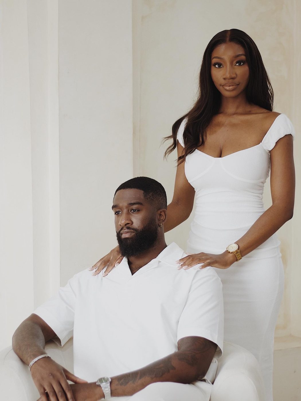 A couple dressed in white, one seated and one standing behind with a hand on their shoulder