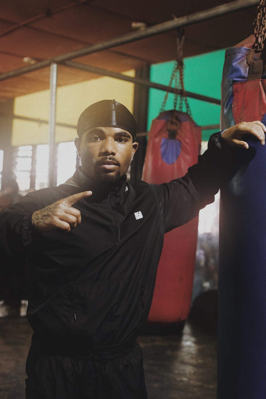 A boxer in a black tracksuit and durag leaning against a punchbag in a gym
